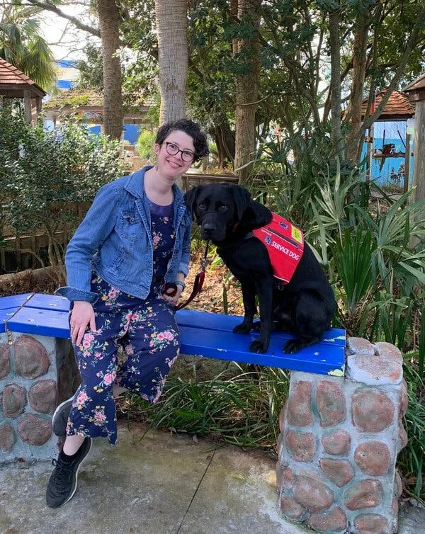 A short-haired woman smiling in a nature park while sitting next to a black service dog held by a leash.